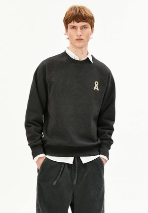 Junger Mann mit kurzen braunen Haaren trägt einen dunkelgrauen Pullover über weißem Hemd und dunkelgrauen Cordhosen, die Hände in den Taschen, neutraler Hintergrund.