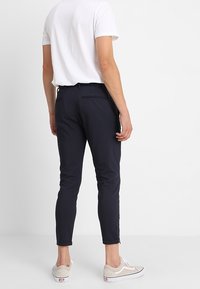 Man som bär slim-fit marinblå byxor, vit kortärmad skjorta och beige sneakers, står vänd mot en vit vägg.