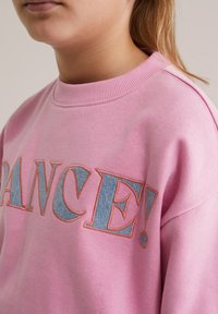 Približek osebe, ki nosi roza pulover z velikimi vezenimi črkami "DANCE!" v modri in oranžni niti.