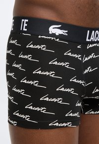 Κοντινή λήψη σε μαύρα εσώρουχα μποξέρ με λευκό μοτίβο κειμένου «Lacoste» και μαύρη μέση με λευκό κείμενο «Lacoste» και λογότυπο κροκόδειλου.