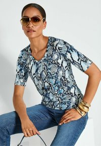 Kurzarmtop mit einem Blau- und Schwarzschlangenmuster, aus weichem Stoff; kombiniert mit blauen Jeans und goldenen Armreifen.