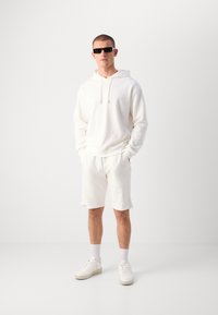 Felpa bianca con logo in rilievo, pantaloni corti bianchi coordinati, sneakers bianche e occhiali da sole scuri. Il materiale sembra texturizzato e informale.
