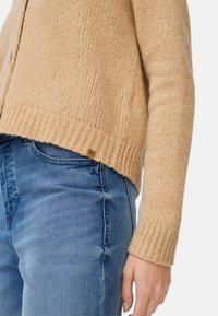 Hellbrauner Strickcardigan mit gerippten Bündchen und Saum, der eine subtile Textur aufweist. Kombiniert mit blauen Jeans. Kleines Markentag sichtbar.