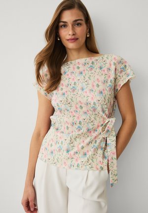 Młoda kobieta z długimi włosami, ubrana w kwiecisty top z krótkim rękawem, zawiązany w talii, oraz białe spodnie na jednolitym tle.