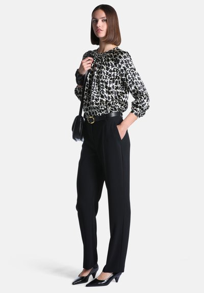 Femme portant un chemisier à motifs noir et blanc, un pantalon noir, une ceinture noire et des chaussures noires à talons bas, tenant un sac à épaule noir.