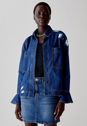 Giacca in denim con chiusura a bottoni, due tasche frontali, dettagli sfrangiati e maniche lunghe; indossata sopra una maglietta nera e una gonna di denim.