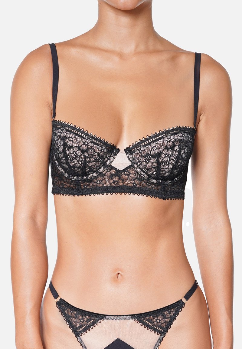 Bralette en dentelle noire avec armature, bords décoratifs festonnés et bretelles ajustables. Le tissu transparent présente des motifs floraux et un panneau nude contrastant.