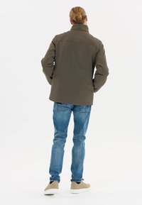 Brun vandtæt jakke med høj krave, parret med lyseblå jeans og beige sko, set fra ryggen mod en hvid baggrund.
