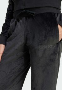 Černé sametové tepláky s jemnou strukturou, které mají nastavitelné stahovací pásky a diskrétní logo adidas na levé straně.