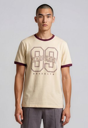 Mann mit kurzen Haaren, der ein beiges T-Shirt mit dunkelrotem Besatz und dem Aufdruck „89 KARL KANI BROOKLYN“ auf der Vorderseite trägt, steht vor einem schlichten Hintergrund.