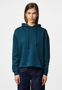 Tealfärgad huvtröja med avslappnad passform, dragsnöre och ribbade ärmmuddar, matchad med mörka jeans. Mjuk textur, avslappnad design.