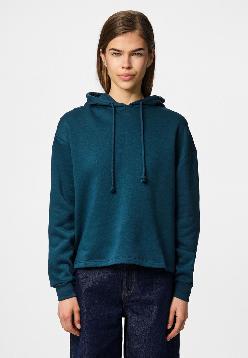 Tealfärgad huvtröja med avslappnad passform, dragsnöre och ribbade ärmmuddar, matchad med mörka jeans. Mjuk textur, avslappnad design.