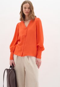 Orange button-up blouse with a V-neck, long balloon sleeves, and textured fabric, paired with light-colored linen trousers and a dark handbag.
