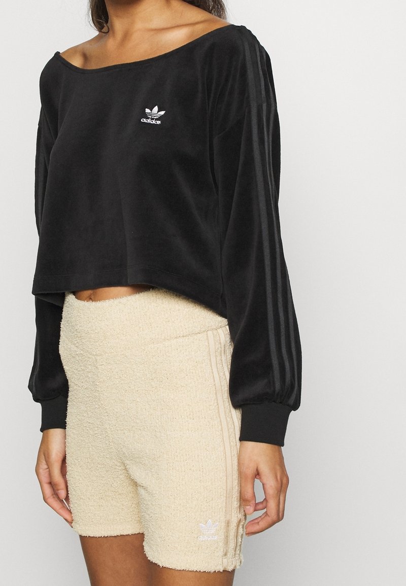 Top corto de terciopelo negro con escote ancho y mangas largas, combinado con pantalones cortos cremosos de textura que presentan un logo blanco y rayas laterales.