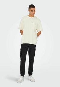 Lys beige oversized T-shirt med en brystlomme og korte ærmer, parret med sorte cargo bukser med sidelommer og elastiske manchetter.