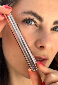 Metallic silver brow serum with a sleek cylindrical shape, clear label, and pink text. Model holds it near their face with manicured nails.