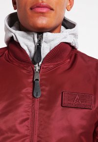 Giacca bombardiere rossa con superficie liscia, colletto a costine e cappuccio grigio. Presenta una chiusura con zip e una toppa con logo sul petto.