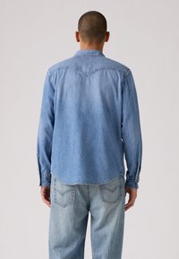 Camisa de mezclilla de color azul claro con un yugo de estilo western, puños abotonados y dobladillo curvado. Usada con jeans de color azul claro a juego, mostrando la vista trasera.