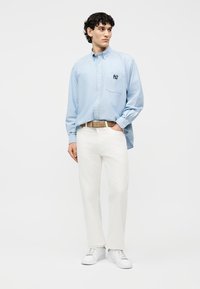 Vyras stovi, vilkėdamas šviesiai mėlyną marškinių su sagomis su logotipu, baltas kelnes, šviesaus atspalvio diržą ir baltus sportinius batelius, žiūrėdamas šiek tiek į dešinę.