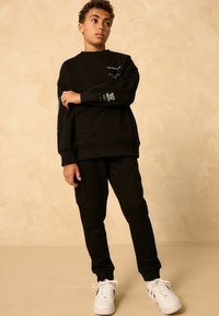 Schwarzes Sweatshirt- und Hosen-Set aus Baumwolle, mit entspannter Passform und bedruckten Akzenten in hellen Farben. Getragen mit weißen Turnschuhen.