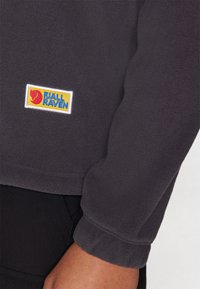 Harmaa fleece-sweatshirt, jossa on ribbaus hihansuissa. Rintataskussa on värikäs Fjällräven-logopatch. Pehmeä tekstuuri ja rento istuvuus.