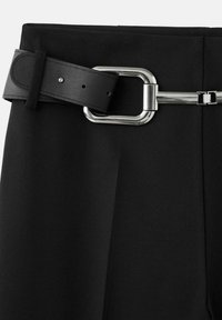 Pantalones negros ajustados con un ancho cinturón de cuero negro que presenta una gran hebilla rectangular plateada y un pequeño detalle metálico.