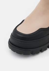 Černá kožená platformová bota s masivní texturovanou podrážkou a hladkou béžovou špicí, která má kontrastní zakřivené švy.
