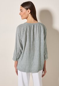 Blusa a righe verde chiaro in lino con maniche a tre quarti, schiena arricciata e polsini con bottoni, abbinata a pantaloni bianchi.