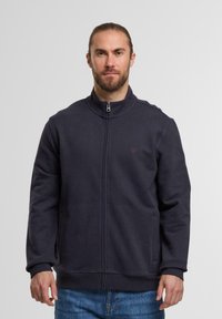 Mann mit Bart und langen, zurückgebundenen Haaren, der eine dunkelblaue Zip-Jacke und blaue Jeans trägt, vor einem schlichten hellen Hintergrund.