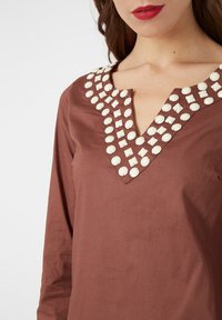 Blusa de algodón marrón con escote en forma de V, decorada con botones circulares blancos a lo largo del cuello. Mangas largas y una textura suave.