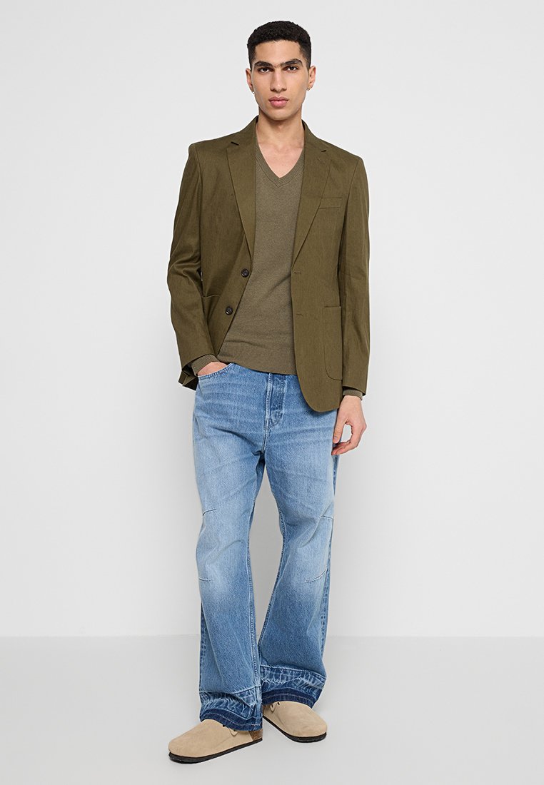 Olivgrön kavaj över en olivgrön V-ringad tröja, parat med ljusblå jeans och beigea slip-on skor. Släta texturer och skräddarsydd passform.