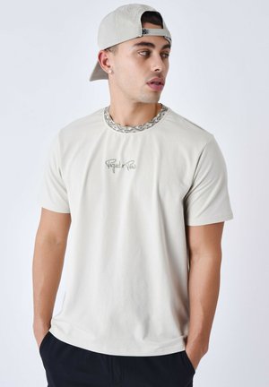Camiseta de manga corta en gris claro con un cuello estampado y el logotipo bordado "Project x Paris". Textura de tela suave, corte relajado.