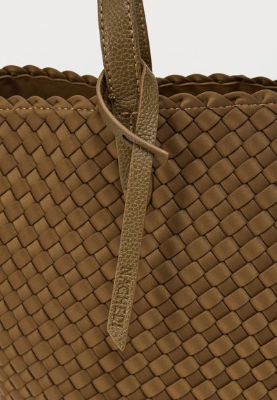 Πλεκτή καφέ τσάντα με υφή στην επιφάνεια, λείες δερμάτινες λεπτομέρειες και λουρί με ετικέτα επωνυμίας. Ο σχεδιασμός είναι δομημένος και ορθογώνιος.