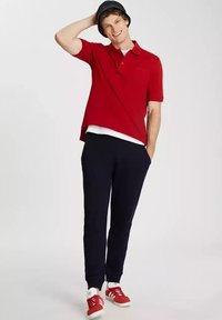 Czerwona koszulka polo z krótkim rękawem, zestawiona z granatowymi spodniami i czerwonymi trampkami. Osoba nosi granatowy kapelusz typu bucket. Ubranie casualowe.