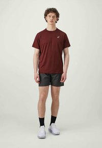 T-shirt courte à manches en bordeaux associée à un short noir. Le modèle porte des baskets blanches et des chaussettes noires. Design simple avec un logo sur la poitrine.