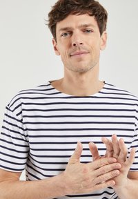 Camiseta blanca con rayas horizontales marinas, cuello redondo, textura suave, con un corte relajado y un diseño casual. Manos juntas.