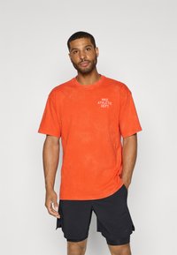 Orange bomulds t-shirt med en afslappet pasform og rund halsudskæring, med hvid tekst "NIKE ATHLETIC DEPT." på brystet. Rynket tekstur.
