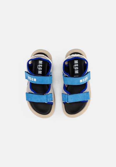 Sandálias azuis e cinzas com tecido em malha, tiras de Velcro ajustáveis, palmilhas pretas e sola em borracha bege. Nome da marca "MSGM" visível.