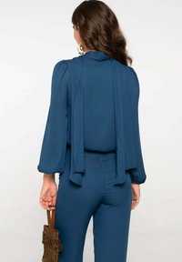 Blusa azul marino de manga larga con detalle de lazo en la parte posterior, combinada con pantalones ajustados a juego. Sosteniendo un bolso con flecos dorados.