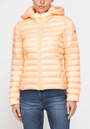 Donna che indossa una giacca imbottita trapuntata arancione chiaro con cappuccio e cerniera, abbinata a jeans di denim blu, in piedi davanti a uno sfondo semplice.
