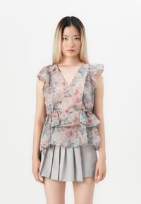 Blusa avvolgente floreale in tessuto trasparente con maniche a volants, caratterizzata da una combinazione di motivi rosa e blu. Abbinata a una gonna plissettata grigia.