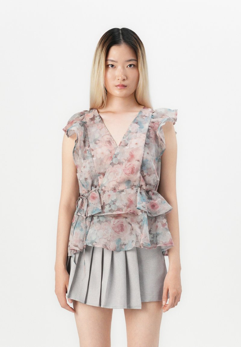 Blusa avvolgente floreale in tessuto trasparente con maniche a volants, caratterizzata da una combinazione di motivi rosa e blu. Abbinata a una gonna plissettata grigia.