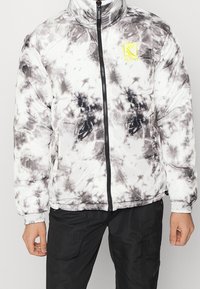 Kurtka puchowa w czarno-białym wzorze tie-dye, z wysokim kołnierzem, zapięciem na zamek oraz żółtym logo na lewej piersi.