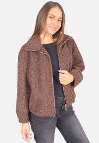 Braune flauschige Zip-Jacke mit hohem Kragen, strukturierter Oberfläche, seitlichen Taschen und lockerer Passform. Getragen über einem schwarzen Oberteil und grauen Hosen.