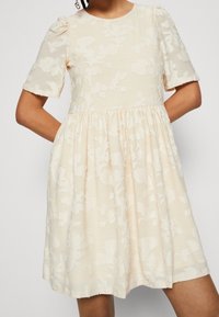 Helles beige Kleid mit kurzen Puffärmeln, strukturiertem Blumenmuster, rundem Ausschnitt und gerafftem Rock. Der Saum ist leicht ausgefranst.