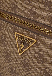 Braune Stofftasche mit Muster, versehen mit einem dreieckigen Logo in Goldton und einem Metallreißverschluss, der die Designdetails hervorhebt.