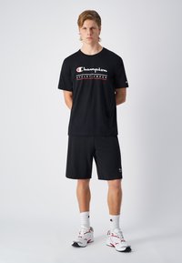 Camiseta atlética negra y pantalones cortos con logo bordado de "Champion Athleticwear" en blanco y rojo. Calcetines blancos y zapatillas deportivas completan el look.