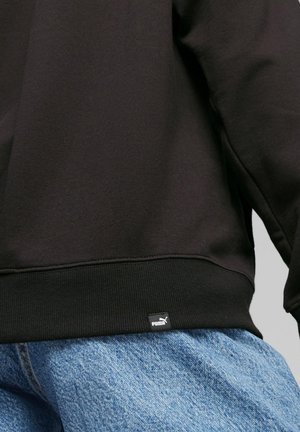 Czarny brzeg bluzy Puma z metką marki noszony na jasnoniebieskich dżinsach na szarym tle.