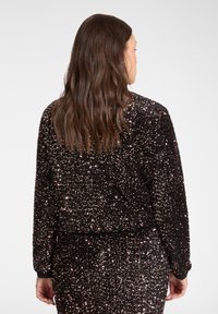 Schwarze Pailletten-Cropped-Jacke mit langen Ärmeln, die eine weiche Textur und einen taillierten Bund aufweist. Die Rückseite zeigt ein schimmerndes Muster.
