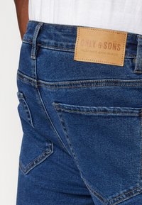 Tmavě modré džíny s koženou visačkou označenou "ONLY & SONS." Obsahují viditelné švy, dvě zadní kapsy a texturovaný denimový povrch.